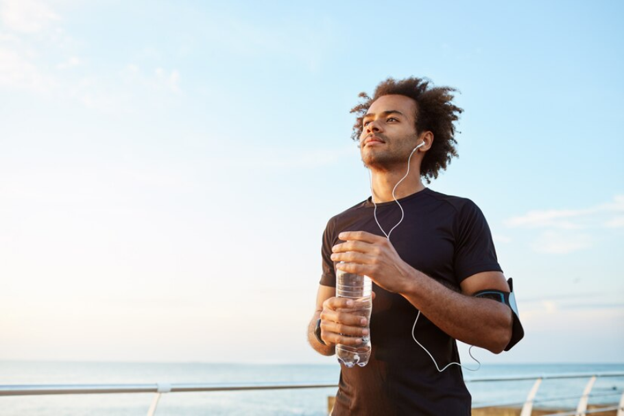 Man athlete drinking water out of plastic bottle after hard running workout. dark-skinned male sportsman looking at the sky while running enjoying view
