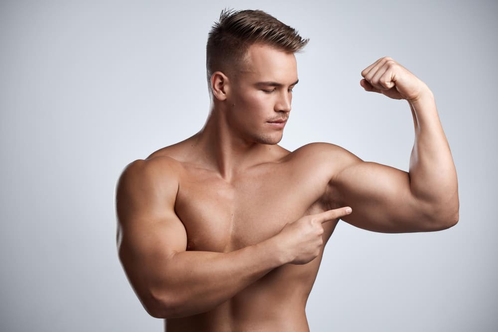 Man bodybuilder and pointing to bicep in studio background and exercise for muscular power strong sexy and topless male model sports athlete and flex arm muscle with pride of fitness results